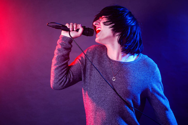 The young guy vocalist is screaming in microphone on stage on concert illuminated with red and blue lights. - Photo, Image