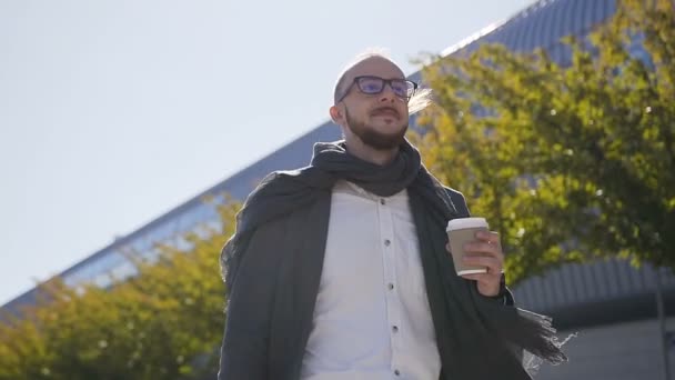 Portrait of the Caucasian handsome businessman in glasses walking in the street near office building , drinking coffee and carrying a suitcase. Outdoors - Video