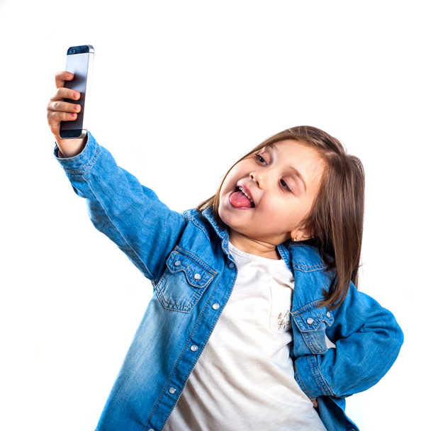 美しい少女は、白い背景の上にスマート フォンを使用して selfie をやっています。. - 写真・画像