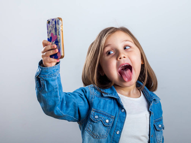Beautiful little girl is doing selfie using a smart phone, on light background. - Foto, afbeelding