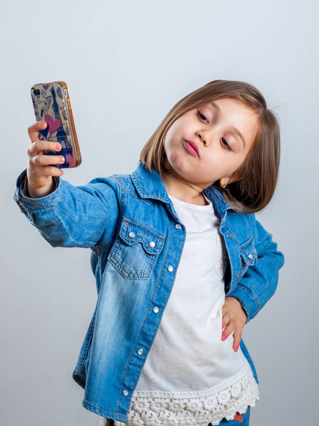 Beautiful little girl is doing selfie using a smart phone, on light background. - Φωτογραφία, εικόνα