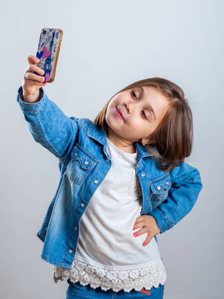 Beautiful little girl is doing selfie using a smart phone, on light background. - Photo, Image