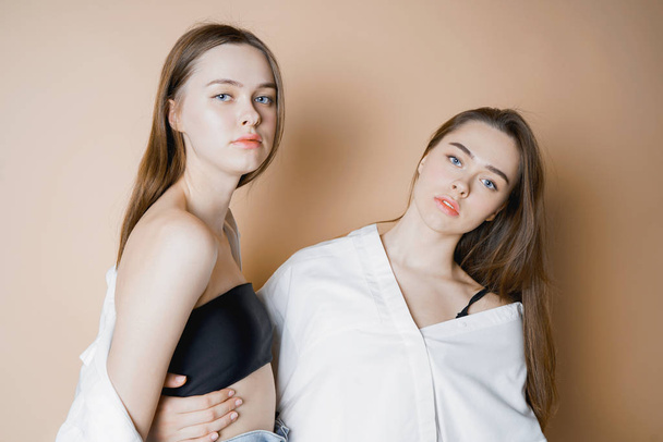 Mody modele dwie siostry bliźniaczki piękne dziewczyny nago, patrząc na kamery na białym tle na beżowym tle                                - Zdjęcie, obraz