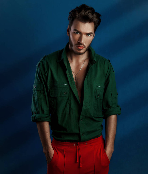 Fashion portrait of young man in green shirt and red pants poses over dark blue wall with contrast light - Фото, изображение