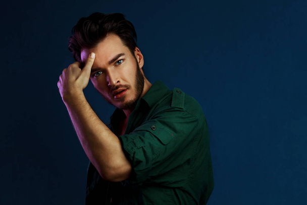 Fashion portrait of young man in green shirt and style haircut poses over dark blue wall with purple contrast color light - Φωτογραφία, εικόνα