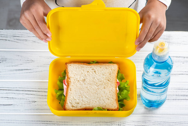 Su un tavolo di legno bianco c'è una scatola da pranzo con pane tostato con prosciutto e lattuga e una bottiglia di plastica d'acqua. Schoolboy tiene con le mani la copertina di un cestino da pranzo
  - Foto, immagini