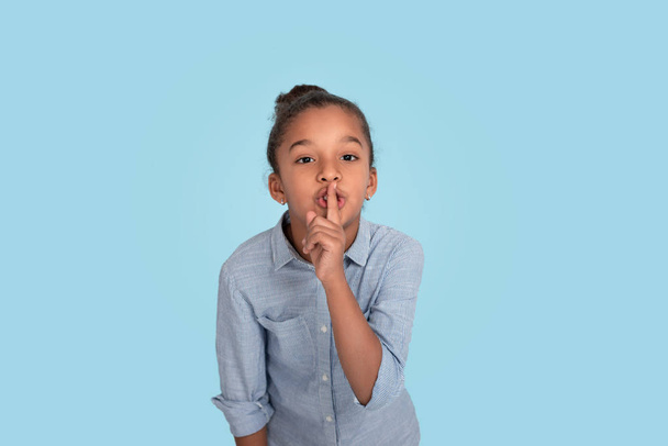 Close up emotional portrait of mulatto frizzy girl wearing blue shirt. She holds her index finger at her lips and asks with this gesture not to talk about her secret. - Foto, Imagen