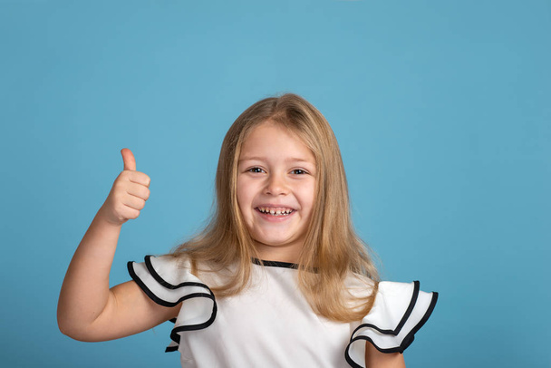 Close up emotional portrait of young blonde  smiling girl wearing white blous with black strips on blue background in studio. She raised her finger up and gestures that everything is fine. - Photo, Image