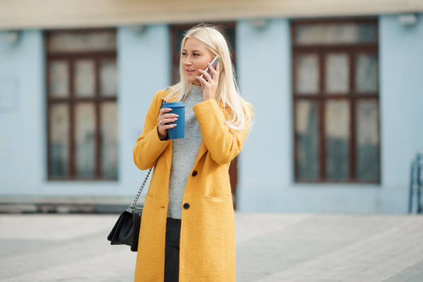 Счастливая молодая блондинка в желтом ярком пальто разговаривает по телефону и ждет встречи друзей. Держит чашку кофе, чтобы пойти
. - Фото, изображение