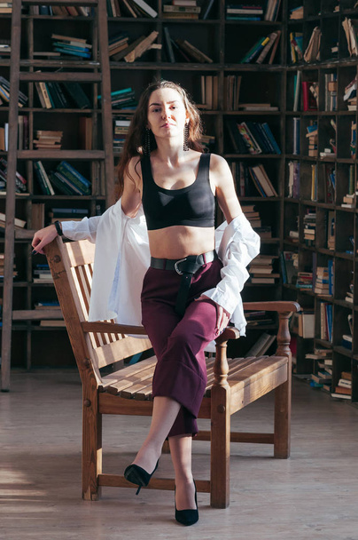 Attractive young female wearing black sport bra lingerie and white shirt unbuttoned, casual fashion style, sitting on wooden chair, bookshelves in the backdrop. Long hair, sensual beautiful seductive - Fotoğraf, Görsel