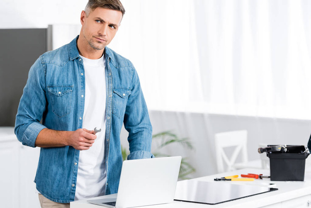 handsome man holding wrench while standing near laptop  - Фото, изображение