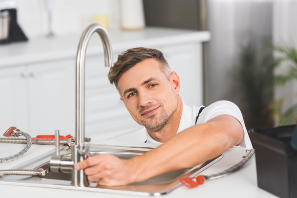 smiling adult repairman with spanner repairing faucet at kitchen and looking at camera - Φωτογραφία, εικόνα
