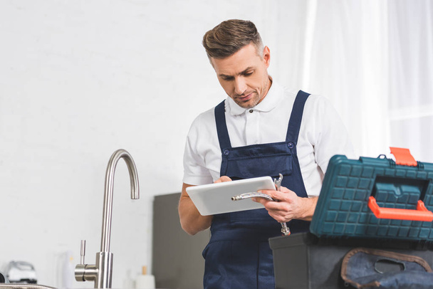 adult repairman using digital tablet and holding tools while repairing faucet at kitchen  - Foto, Imagem