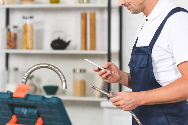 cropped view of adult repairman holding pipe for kitchen faucet repairing and using smartphone - Φωτογραφία, εικόνα