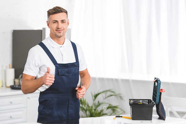 adult repairman holding screwdriver and showing thumb up sign at kitchen and looking at camera  - Φωτογραφία, εικόνα