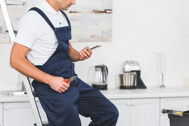 cropped view of repairman sitting on ladder holding pliers and using smartphone at kitchen  - Photo, Image