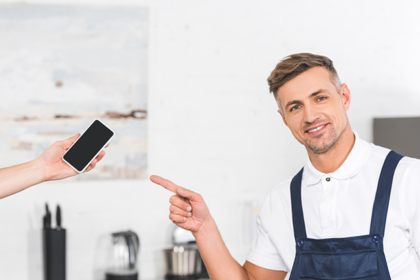 partial view of hand holding smartphone and smiling adult repairman pointing at blank screen  - Fotografie, Obrázek