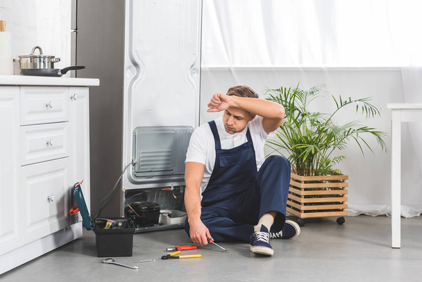 tired adult repairman sitting on floor with hand on forehead and looking at tools while repairing refrigerator    - Foto, Bild