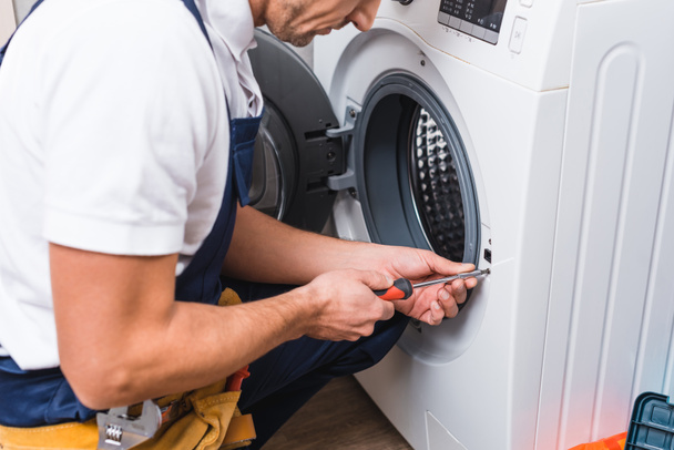 Ausgeschnittene Ansicht eines erwachsenen Reparateurs, der mit einem Schraubenzieher arbeitet, während er Waschmaschine im Badezimmer repariert  - Foto, Bild