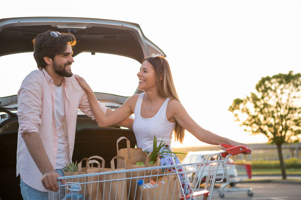 Ένα ευτυχισμένο ζευγάρι βάζει παντοπωλεία στο αυτοκίνητό του μπροστά από ένα σούπερ μάρκετ. - Φωτογραφία, εικόνα