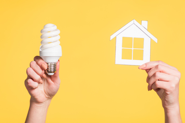 bijgesneden beeld van vrouwelijke handen met papier huis en fluorescentielamp in handen geïsoleerd op geel, energie-efficiëntie thuis concept - Foto, afbeelding