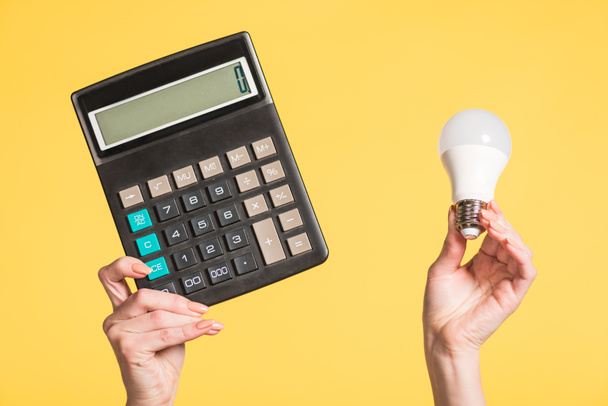 cropped view of woman holding fluorescent lamp and calculator in hands isolated on yellow, energy efficiency concept  - Photo, Image