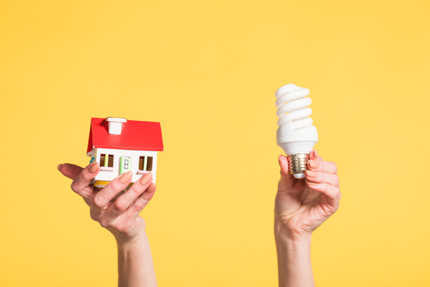 cropped view of woman holding fluorescent lamp and house model in hands isolated on yellow, energy efficiency at home concept - Photo, Image