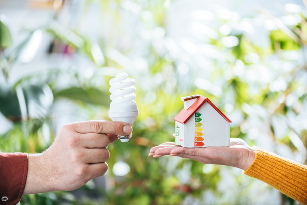 vista recortada del hombre sosteniendo lámpara fluorescente cerca de la mujer con la casa de cartón en la mano, concepto de eficiencia energética en el hogar
 - Foto, imagen