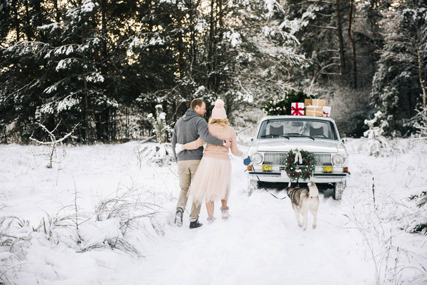A guy and a girl getting ready for Christmas, walking the dog husky on a background of vintage car, on the roof tree and gifts in the winter snowy forest - Fotografie, Obrázek
