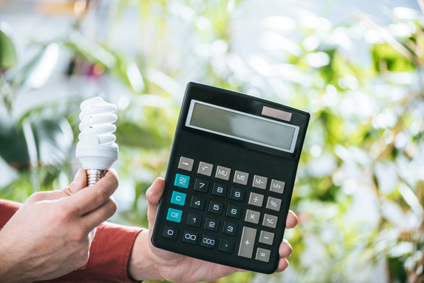 cropped view of fluorescent lamp and calculator in male hands, energy efficiency concept - Photo, Image