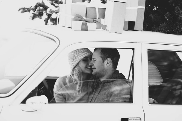A guy and a girl ride in a retro car decorated with a Christmas tree and presents in a snowy forest. Black and white photography - Photo, image