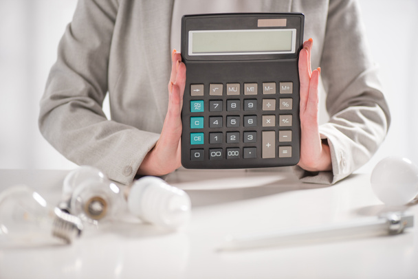 cropped view of woman holding calculator near lamps on white background, energy efficiency concept - Photo, Image