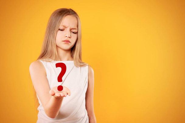 Pensive teen girl holding question mark over yellow background. Problem concept. Blonde girl thinking  - Photo, Image