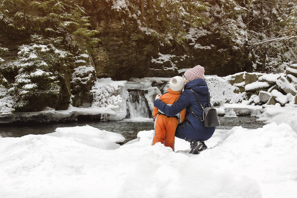 Anya és fia ül a háttérben a hóval borított vízesés, hegy- és erdeifenyő erdők ölelésében. Téli felhős nap - Fotó, kép