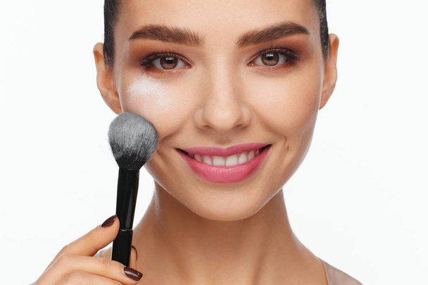 Portrait of a beautiful smiling young woman holding a brush in her hand and applying powder to her face with a brush. White background - Valokuva, kuva