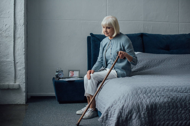 lonely pensive senior woman sitting on bed with walking stick at home - Fotoğraf, Görsel