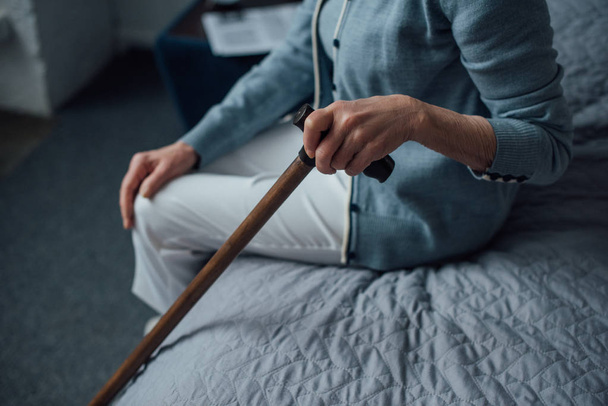 cropped view of senior woman sitting on bed and holding walking stick at home - Foto, Bild