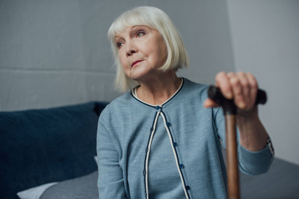 lonely senior woman sitting on bed with walking stick at home - Фото, изображение