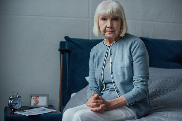 Starší žena při pohledu na fotoaparát a sedí na posteli s sepjatýma rukama doma - Fotografie, Obrázek
