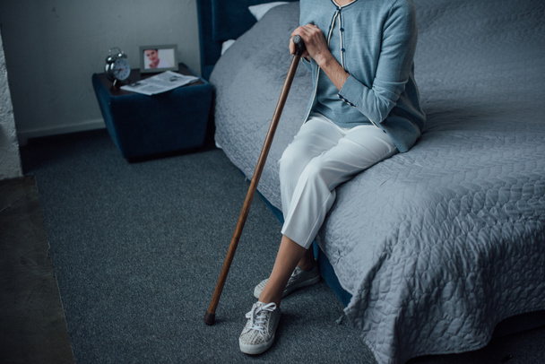 częściowy widok starszy kobieta siedzi na łóżku z laską w domu - Zdjęcie, obraz