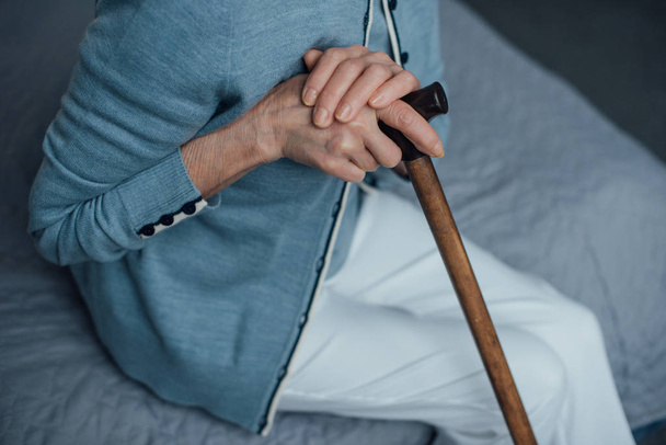 cropped view of senior woman sitting on bed with walking stick at home - Fotó, kép