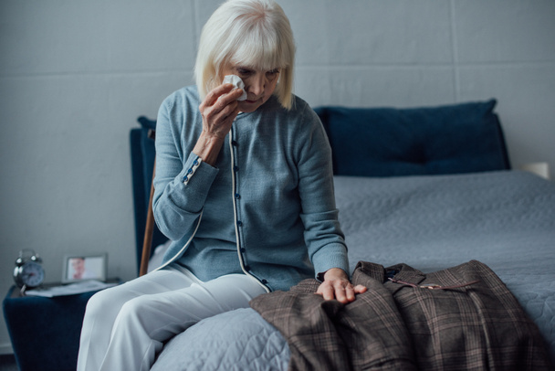Selektywny fokus płacz starszy kobieta siedzi na łóżku, ocierając łzy i ogląda kurtka w domu - Zdjęcie, obraz