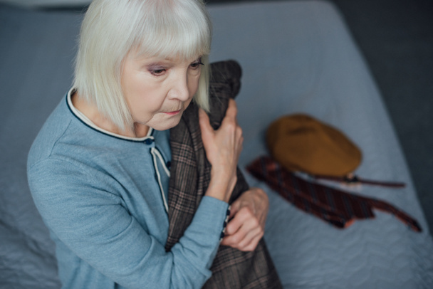 αναστατωμένος ανώτερων γυναίκα με γκρίζα μαλλιά, κάθεται στο κρεβάτι και κρατώντας το σακάκι στο σπίτι - Φωτογραφία, εικόνα