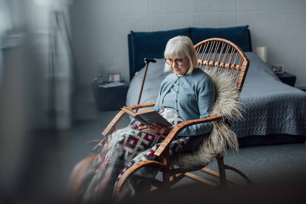 Senior vrouw zitten in rieten schommelstoel en leesboek in slaapkamer - Foto, afbeelding