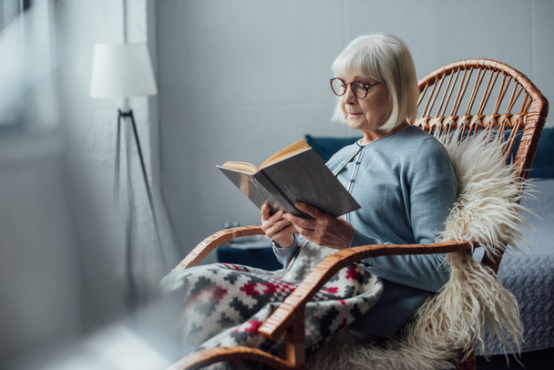 senior woman in glasses sitting in wicker rocking chair and reading book at home - Photo, Image