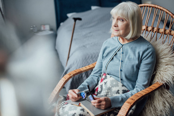 zamyšlený starší žena seděla v houpacím křesle s knihou doma proutí - Fotografie, Obrázek