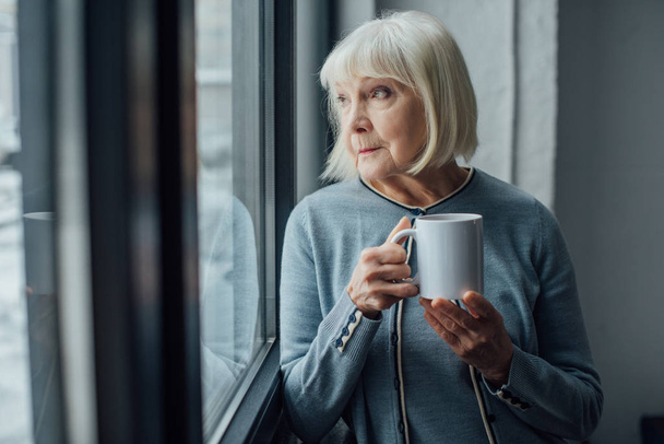 mujer mayor sentada cerca de la ventana y bebiendo café en casa
  - Foto, Imagen