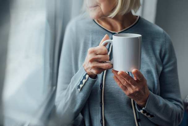 обрезанный вид пожилой женщины, держащей чашку кофе дома
  - Фото, изображение