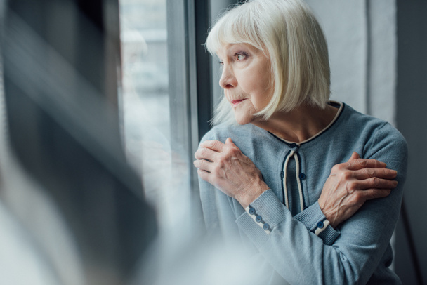 thoughtful senior woman with hands crossed looking through window at home  - Foto, Bild
