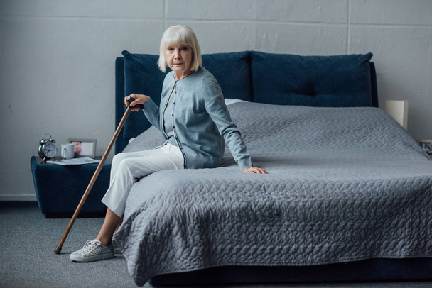 senior woman sitting on bed with walking stick at home and looking at camera - Foto, Imagen